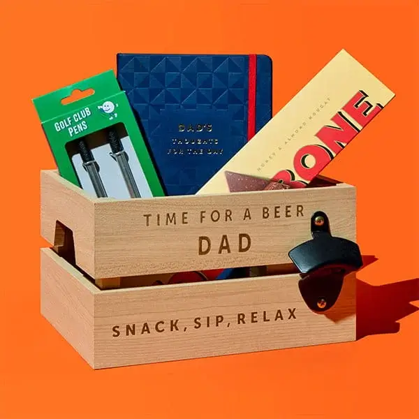 A wooden gift crate engraved with Time for a Beer Dad and Snack Sip Relax, filled with a Toblerone bar, golf club pens, a blue notebook and small gifts, displayed against a bright orange background.